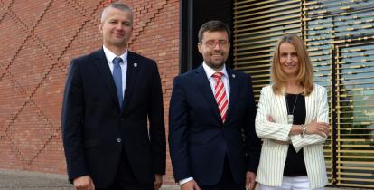 Na zdjęciu trzech uśmiechniętych przedstawicieli samorządu gminy Konstancin-Jeziorna pozuje przed nowoczesnym budynkiem z ceglaną i przeszkloną elewacją.