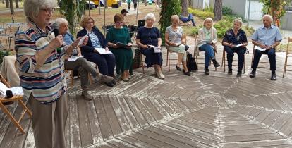 Grupa osób siedzi w altanie, jedna osoba czyta książkę.
