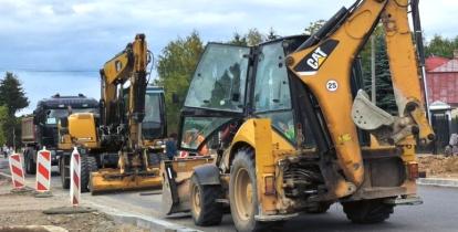 Maszyny budowlane prowadzące prace przy przebudowie drogi, z ułożonym już fragmentem nowego asfaltu i krawężnikami.