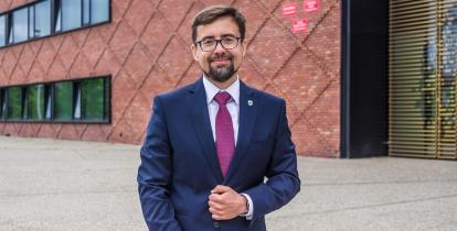 uśmiechnięty burmistrz w granatowym garniturze stoi na tle urzędu miasta i gminy Konstancin-Jeziorna