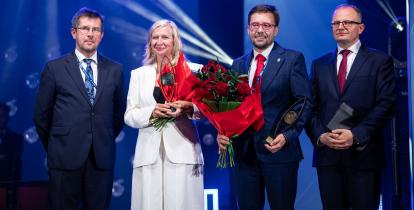 Trzy osoby stoją na scenie, na niebieskim tle. 