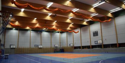 Wnętrze dużej, nowoczesnej hali sportowej z wieloma koszami do koszykówki, niebieską podłogą i pomarańczowymi siatkami pod sufitem.