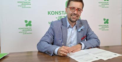 Uśmiechnięty mężczyzna w okularach i eleganckiej marynarce siedzi przy biurku i podpisuje dokumenty na tle banneru promocyjnego miasta.