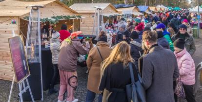 Ludzie spacerują między drewnianymi stoiskami z ozdobami i świątecznymi produktami podczas jarmarku w parku otoczonym drzewami.