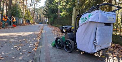 Odkurzacz do liści na ulicy zasypanej liściami, w tle dwoje pracowników z grabiami porządkuje liście.