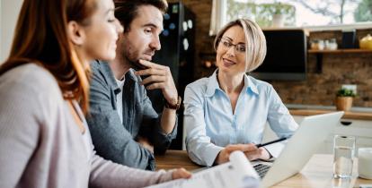 Trzy osoby siedzą przed komputerem, na stole leżą dokumenty, sprawdzają coś