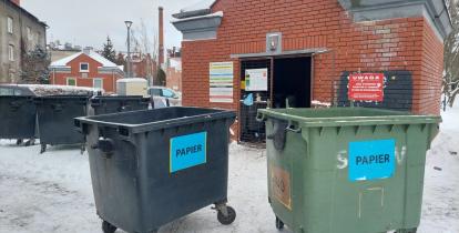 Altana śmietnikowa na osiedlu zimą, przed którą stoją przepełnione pojemniki na papier, ustawione na zaśnieżonym placu.