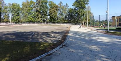 Szkolne boisko z asfaltową nawierzchnią i bramkami, położone obok budynku szkoły, otoczone zielenią i oświetleniem.