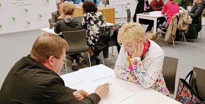 Mieszkańcy uczestniczą w warsztatach konsultacyjnych w sali urzędu, którzy przy stolikach omawiają mapy i dokumenty z przedstawicielami gminy.