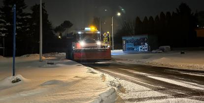 Pług śnieżny odśnieżający zaśnieżoną jezdnię, oświetloną latarniami ulicznymi, z widocznymi śladami przejazdu na mokrym asfalcie.