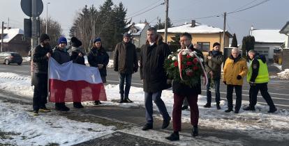Grupa osób podczas zimowej uroczystości składa kwiaty pod pomnikiem, jedna z nich niesie wieniec, a w tle widoczna jest flaga Polski oraz zabudowa jednorodzinna przy drodze.