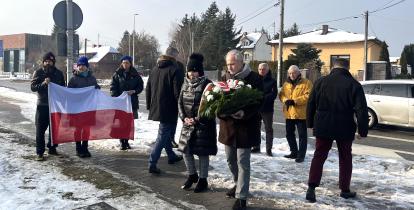 Grupa osób podczas zimowej uroczystości składa kwiaty pod pomnikiem, jedna z nich niesie wieniec, a w tle widoczna jest flaga Polski oraz zabudowa jednorodzinna przy drodze.