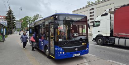 Niebieski autobus komunikacji miejskiej linii L55 stoi na przystanku, do którego wsiadają pasażerowie, a obok na jezdni przejeżdża samochód ciężarowy.