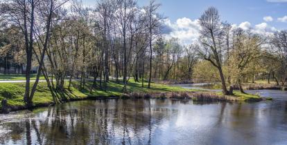 Parkowy krajobraz z rzeką, zielonymi trawnikami i drzewami odbijającymi się w spokojnej wodzie pod błękitnym, częściowo zachmurzonym niebem.