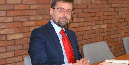 Mężczyzna w granatowym garniturze i czerwonym krawacie siedzi przy stole konferencyjnym, trzymając dłonie złożone na blacie, na tle ceglanej ściany.