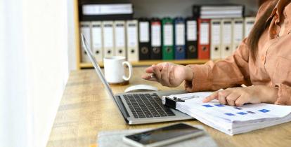 Kobieta siedzi przy biurku i analizuje dokumenty finansowe, pracując na laptopie w biurze z segregatorami na półkach w tle.