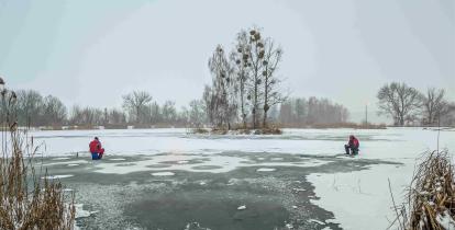 Dwóch wędkarzy siedzi na zamarzniętym stawie i łowi ryby w przeręblach, mimo cienkiej i nierównej tafli lodu pokrytej śniegiem.