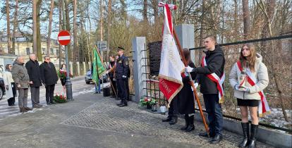Grupa młodych dziewczyn i chłopców z biało-czerwonymi wieńcami i szarfami oraz trzech starszych mężczyzn i trzy kobiety stoją naprzeciwko tablicy zawieszonej na ogrodzeniu. Przy ogrodzeniu dwóch strażników miejskich w galowych mundurach.
