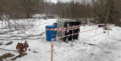 Na zaśnieżonym terenie przy zadrzewionej drodze stoją zabezpieczone taśmą ostrzegawczą pojemniki – mauzer i dwie beczki – odgrodzone drewnianymi palikami.