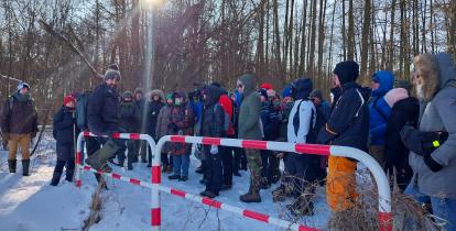 Grupa kilkudziesięciu osób w zimowych kurtkach i czapkach stoi na zaśnieżonej leśnej polanie podczas spaceru przyrodniczego, słuchając przewodnika, który opowiada o terenie mokradeł w mroźny, słoneczny dzień.