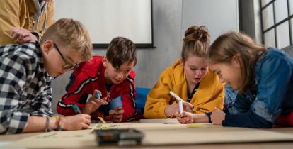 Grupa dzieci siedzi przy stole i wspólnie rysuje na dużych arkuszach papieru, korzystając z kolorowych markerów, pod opieką dorosłego opiekuna w sali zajęć.