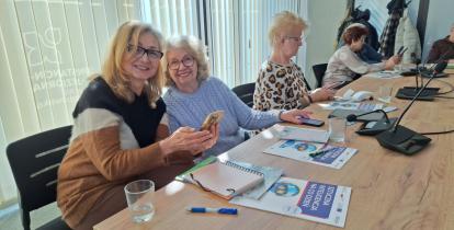 Dwie kobiety siedzą przy stole, patrzą w obietkyw i uśmiechają się. W rękach trzymają telefony komórkowe, przed nimi na stole leżą broszury, zeszyty - są na szkoleniu.