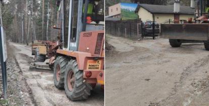 Równiarka i sprzęt drogowy prowadzą prace przy wyrównywaniu i utwardzaniu nawierzchni leśnego duktu wśród zabudowy i drzew.