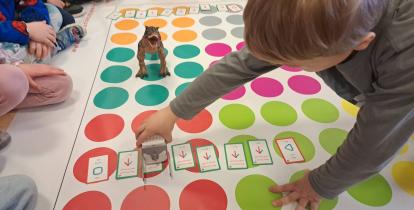 Grupa dzieci w przedszkolu siedzi na dywanie wokół dużej maty edukacyjnej z kolorowymi polami. Chłopiec na pierwszym planie ustawia małego robota oraz karty z komendami ruchu (strzałkami), planując drogę do figurki dinozaura.