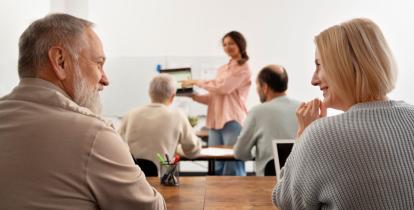 Widok zza pleców pary seniorów siedzących przy biurku w sali wykładowej. Mężczyzna z brodą i kobieta o jasnych włosach uśmiechają się do siebie. Na pierwszym planie widać organizer z przyborami do pisania. W głębi sali, na rozmytym tle, inna uczestniczka prowadzi prezentację.