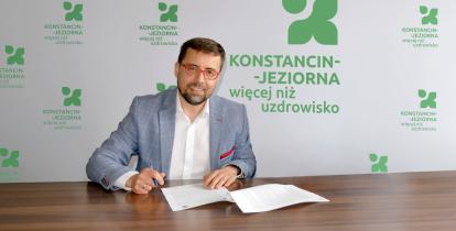 Mężczyzna w okularach podpisuje dokumenty przy stole na tle ścianki z logo „Konstancin-Jeziorna – więcej niż uzdrowisko”.