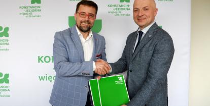 Dwóch mężczyzn w garniturach uściskiem dłoni potwierdza podpisanie dokumentu na tle ścianki z logo Konstancina-Jeziorny.