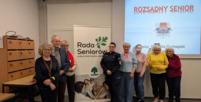 Grópa seniorów stoi w sali przy roll-upie z napisem Rada Seniorów, w tle na ścianie slajd prezentacji Rozsądny Senior