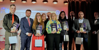 Grupowe zdjęcie dziesięciu osób na podświetlonej scenie, uchwycone podczas ceremonii wręczania nagród „Perła Mazowsza 2024”. W pierwszym rzędzie laureaci trzymają dyplomy i papierowe torby z nagrodami.