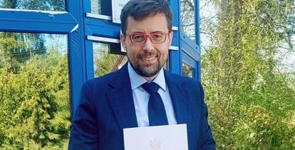 Mężczyzna w garniturze stoi przed budynkiem i trzyma w rękach dokument zatytułowany „Akt notarialny”.