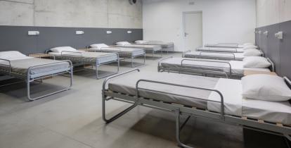 Nowoczesne pomieszczenie schronowe z betonowymi ścianami i oświetleniem LED, w którym ustawiono rzędy metalowych łóżek z materacami i poduszkami.