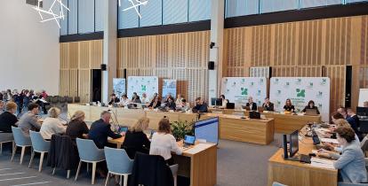 Grupa osób siedzi za stołami na sali posiedzeń.