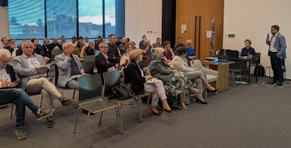 Uczestnicy spotkania siedzą na sali konferencyjnej i słuchają mężczyzny prezentującego temat przy użyciu mikrofonu i projektora.