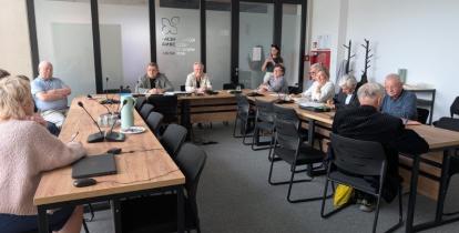 Fotografia przedstawia posiedzenie Rady Seniorów Gminy Konstancin-Jeziorna w nowoczesnej sali konferencyjnej. Grupa kilkunastu osób siedzi przy stołach ułożonych w kształt litery U. Na pierwszym planie po lewej widać tył sylwetki kobiety w różowym swetrze, a przed nią laptop i mikrofon konferencyjny na drewnianym blacie. Pozostali uczestnicy obrad, w większości seniorzy, siedzą przodem lub bokiem do obiektywu, słuchając wystąpienia. W głębi sali, za szklaną ścianą z drzwiami, na której widnieje logo gminy (