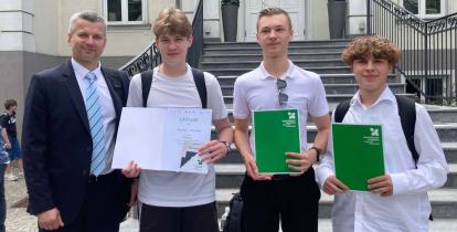 Na schodach przed budynkiem stoją cztery osoby: zastępca burmistrza gminy oraz trzej uczniowie trzymający dyplom i zielone teczki z nagrodami, laureaci konkursu wiedzy o historii Konstancina-Jeziorny.