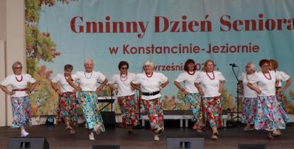Grupa seniorek w jednakowych białych bluzkach, czerwonych koralach i kolorowych, wzorzystych spódnicach tańczy na scenie amfiteatru. W tle widać duży baner z napisem „Gminny Dzień Seniora w Konstancinie-Jeziornie”.