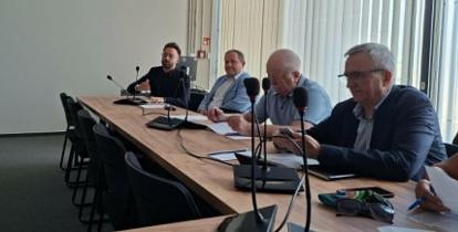 Fotografia przedstawia posiedzenie Rady Seniorów Gminy Konstancin-Jeziorna w nowoczesnej sali konferencyjnej. Grupa kilkunastu osób siedzi przy stołach ułożonych w kształt litery U. Na pierwszym planie po lewej widać tył sylwetki kobiety w różowym swetrze, a przed nią laptop i mikrofon konferencyjny na drewnianym blacie. Pozostali uczestnicy obrad, w większości seniorzy, siedzą przodem lub bokiem do obiektywu, słuchając wystąpienia. W głębi sali, za szklaną ścianą z drzwiami, na której widnieje logo gminy (