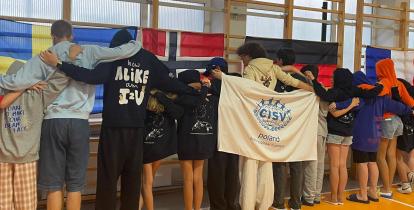 Grupa młodzieży stoi tyłem w szpalerze, obejmując się ramionami w hali sportowej. Przed nimi powieszona jest biała flaga z logo organizacji oraz napisem „CISV poland building global friendship”. W tle na szkolnych drabinkach wiszą flagi państwowe, m.in. Szwecji, Norwegii, Niemiec i Francji.