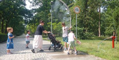 Na parkowej alei kobieta z dziećmi korzysta z bramki zraszającej, która rozpylając wodną mgiełkę przynosi ochłodę podczas upalnego dnia.