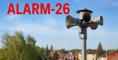 Syrena alarmowa na tle zabudowy i drzew, z czerwonym napisem „ALARM-26”.