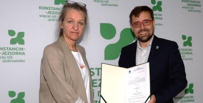 Na zdjęciu kobieta (dyrektor Ewa Winek) i mężczyzna (burmistrz Michał Wiśniewski) stoją przed ścianką z logo gminy Konstancin-Jeziorna. Uściskiem dłoni potwierdzają wręczenie aktu powierzenia stanowiska dyrektora. Mężczyzna trzyma otwartą teczkę z widocznym dokumentem i uśmiecha się do obiektywu.