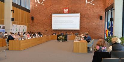 Grupa osób siedzi za stołami na sali posiedzeń