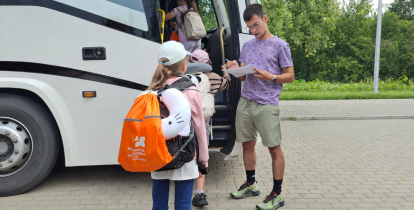 Dzieci z plecakami wsiadają do autokaru przed wyjazdem na wakacyjny wypoczynek w Zakopanem. Opiekun sprawdza listę uczestników.