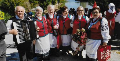 Pięć seniorek w strojach ludowych pozuje do zdjęcia nad brzegiem Wisły. Towarzyszy im mężczyzna trzymający akordeon, a jedna z kobiet ma w rękach wieniec z kwiatów.