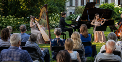Plenerowy koncert muzyki klasycznej w ogrodzie przed zabytkową willą. Muzycy grają na harfie, fortepianie, skrzypcach i wiolonczeli, a publiczność słucha ich, siedząc na krzesłach ustawionych na trawniku