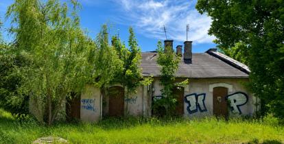 Zaniedbany budynek dawnej parowozowni z zamurowanymi oknami i graffiti na ścianach. Obiekt otaczają wysoka trawa i drzewa